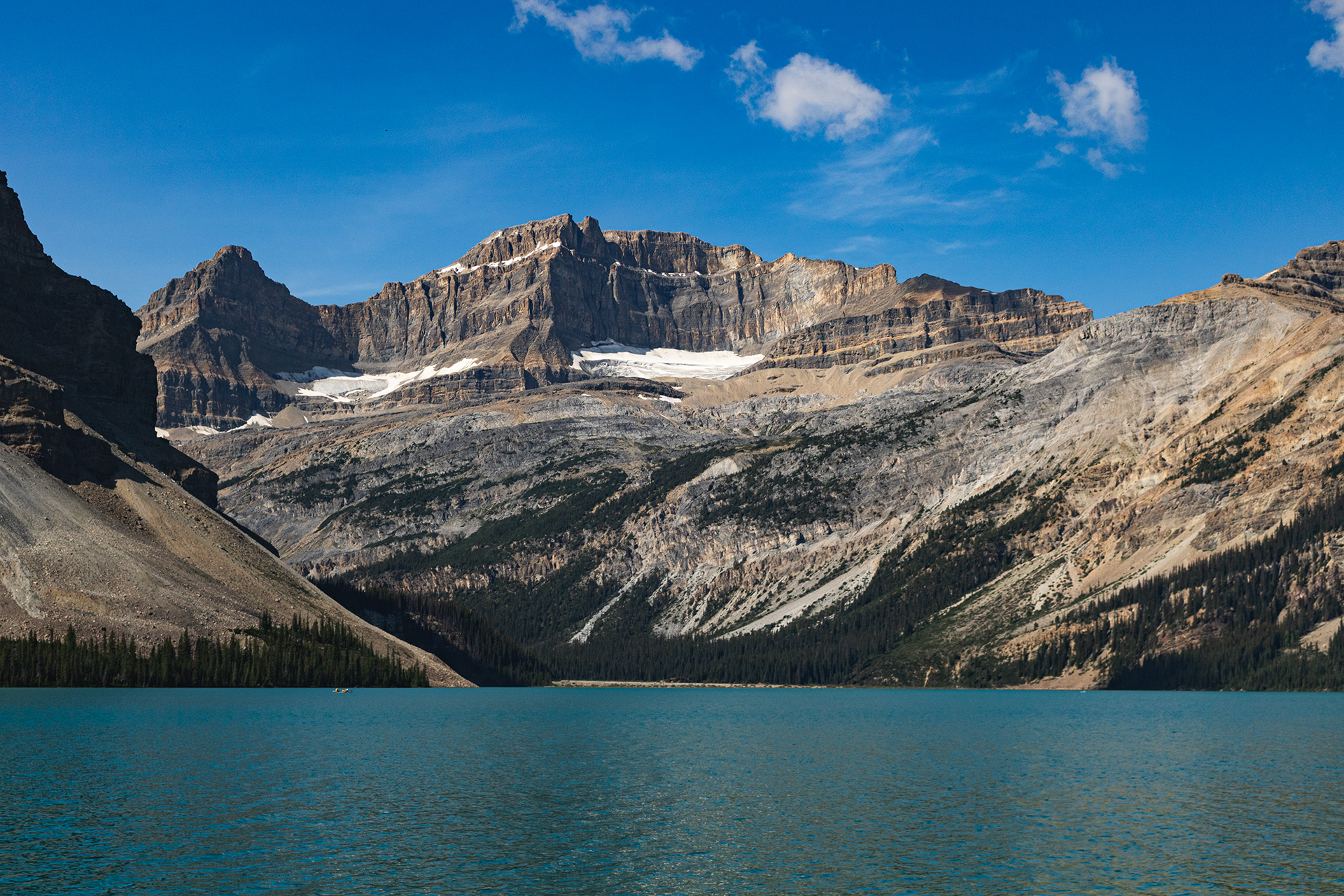 Lake with huge rocky mountain behind it with bits of snow on a plateau halfway up.